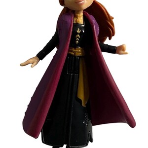 May include: A small plastic figurine of Anna from the Disney film Frozen. She is wearing a black dress with a purple cape, a gold belt, and a gold necklace. Her hair is red and pulled back in a ponytail.