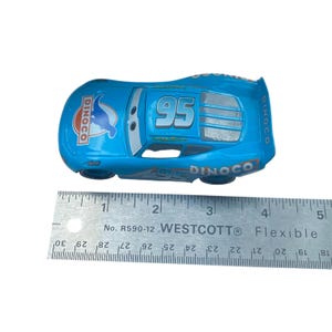 May include: A blue die-cast toy car with the number 95 on the roof and Dinoco branding. The car is approximately 5 inches long and has black wheels. A flexible ruler is placed next to the car for scale.
