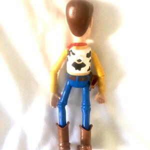May include: A plastic toy figure of Woody from the Toy Story franchise. The figure is brown, yellow, and blue. Woody is wearing a brown cowboy hat, a yellow shirt with a red bandana, and blue jeans. He is holding a brown holster on his right hip.