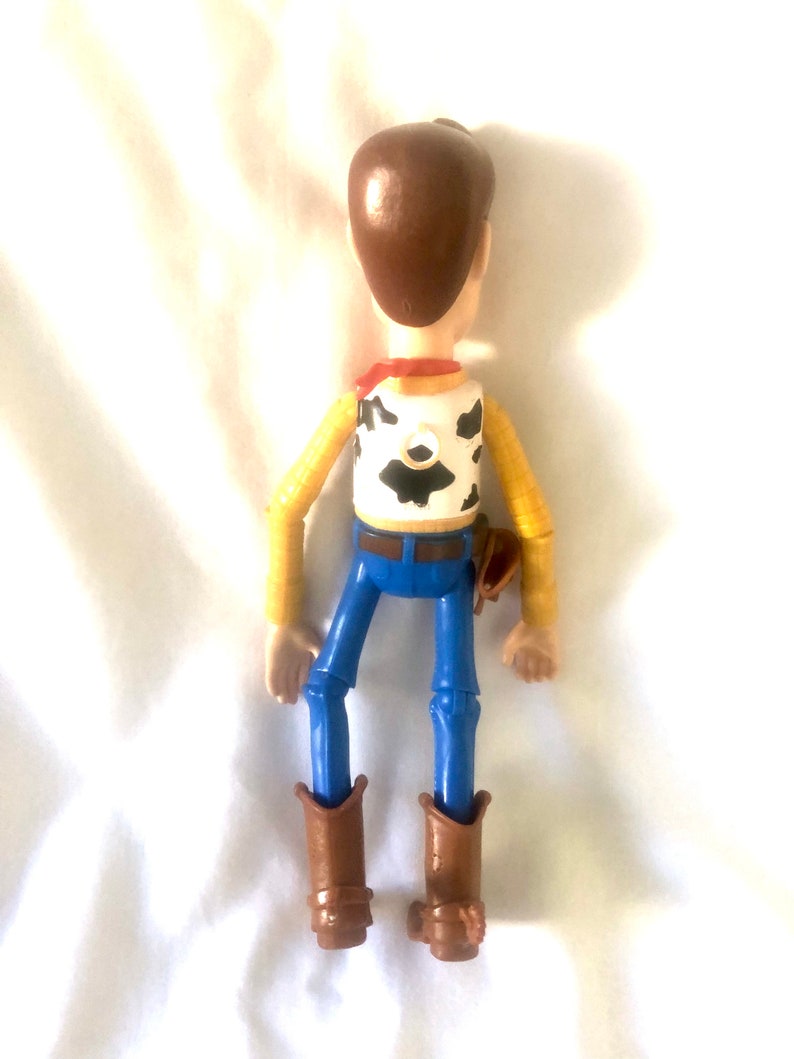 May include: A plastic toy figure of Woody from the Toy Story franchise. The figure is brown, yellow, and blue. Woody is wearing a brown cowboy hat, a yellow shirt with a red bandana, and blue jeans. He is holding a brown holster on his right hip.
