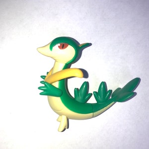 Peut inclure: Figurine en plastique verte et jaune de Sceptile, un Pokémon de type plante. La figurine a une longue queue et un corps vert avec une bande jaune.