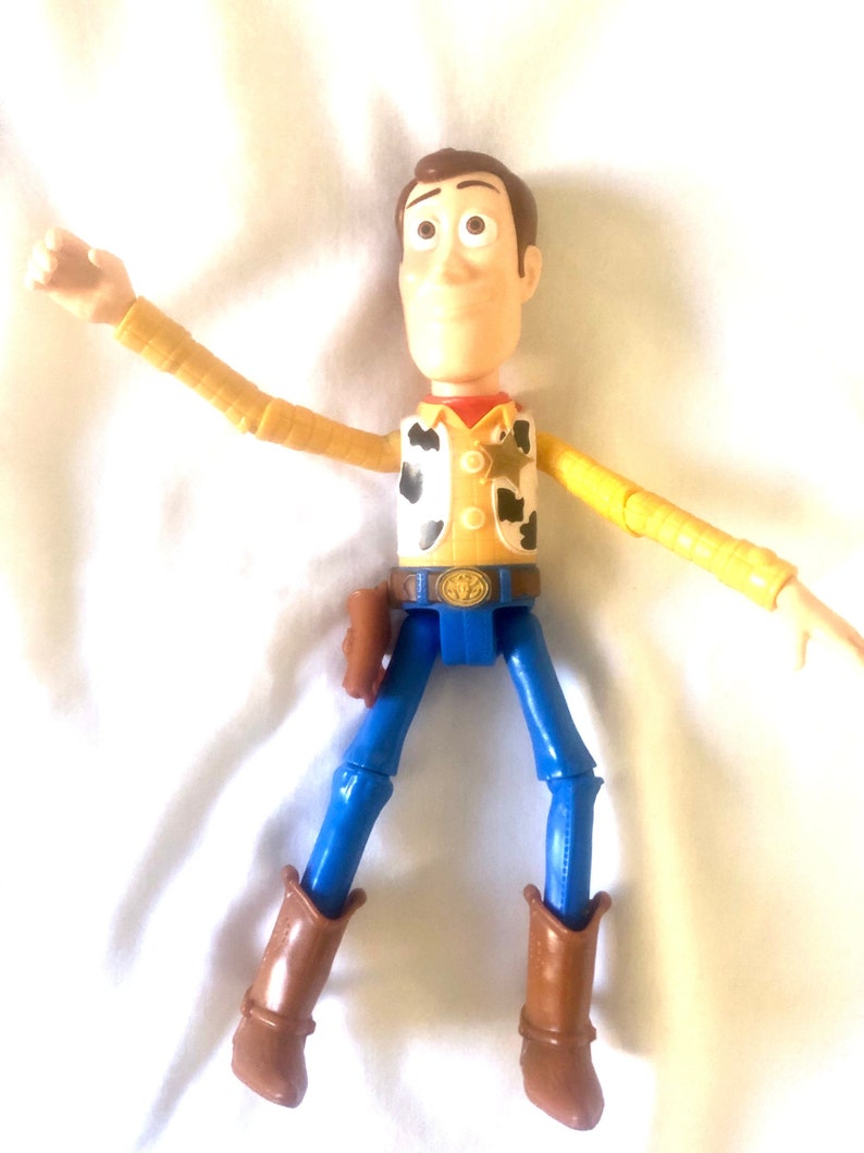 May include: A yellow and brown plastic toy figure of Woody from the Toy Story franchise. Woody is wearing a brown cowboy hat, a yellow shirt with a brown vest, blue jeans, and brown cowboy boots. He has a sheriff's star on his vest and a holster on his hip.