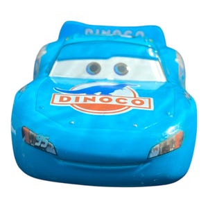 May include: A light blue toy car with the word "DINCO" in a red and white logo on the hood. The car has the number "95" on the side and "DINOSA" on the top. The car has large eyes and a smiling face.