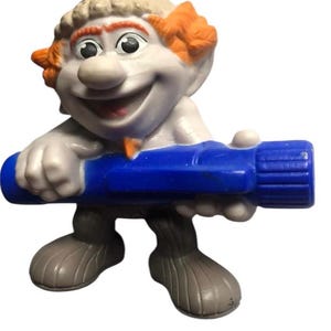 Może przedstawiać: Plastikowa figurka zabawki w kolorze niebieskim i białym przedstawiająca postać z pomarańczowymi włosami, białym kapeluszem i niebieską rurką w ręku.