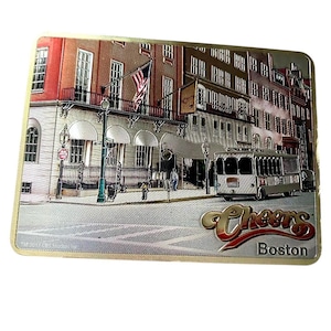 May include: A silver and gold metal sign with a picture of a red brick building with a trolley in front of it. The sign says "Cheers Boston" in gold letters.