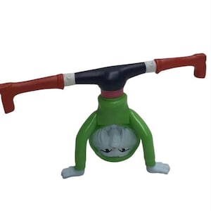 May include: A novelty figurine depicting a character in a handstand. The figure has a green body, a black upper torso, and red arms and feet. The face is white with black eyes and a small mouth. The background is plain white.