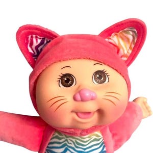 Peut inclure: Un jouet en peluche rose en forme de chat avec un imprimé zèbre arc-en-ciel sur la poitrine et les oreilles. Le jouet a des yeux bruns, un nez rose et un grand sourire.