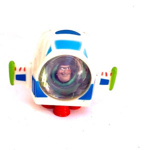 Op de afbeelding: Een wit en blauw speelgoed ruimteschip met Buzz Lightyear uit Toy Story. Het speelgoed heeft rode stuwraketten, groene vleugeldetails en een heldere cockpit met het gezicht van het personage.