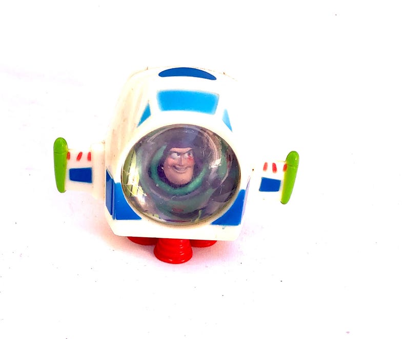 Op de afbeelding: Een wit en blauw speelgoed ruimteschip met Buzz Lightyear uit Toy Story. Het speelgoed heeft rode stuwraketten, groene vleugeldetails en een heldere cockpit met het gezicht van het personage.