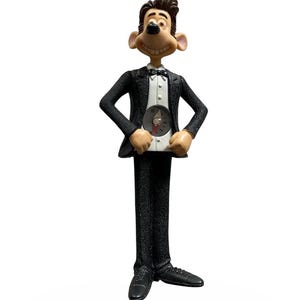 May include: A figurine of a cartoon character in a black tuxedo with a white shirt and bow tie. The character is holding a clock with a red hand. The figurine has a smiling face and is standing on a white background.