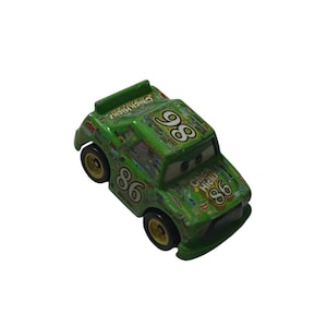 Puede incluir: Coche de carreras de juguete verde con el número 86 y las palabras "Chicki Hicks" impresas en el lateral. El coche tiene ruedas doradas y un alerón. El coche es un juguete de la película Cars.