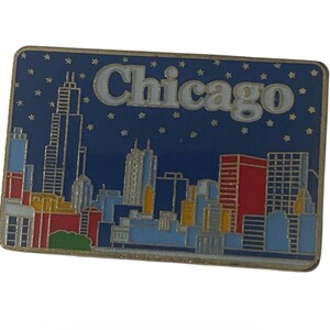 Puede incluir: Un pin rectangular de recuerdo de Chicago con un fondo azul y un borde dorado. El pin presenta un horizonte con la palabra "Chicago" en blanco. Los edificios son de varios colores, incluyendo rojo, azul y amarillo, con pequeños acentos de estrellas.