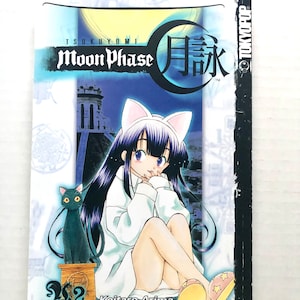 May include: Volume 2 of the Moon Phase manga series by Tsukuyomi, featuring a black cat and a girl with cat ears wearing a white dress and slippers. The cover is blue and white with the title "Moon Phase" in Japanese characters.