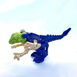 Puede incluir: Un dinosaurio de plástico azul y verde con detalles dorados. El dinosaurio tiene una cola larga y está en posición de carrera.
