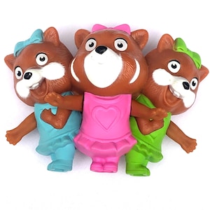 Puede incluir: Tres figuras de juguete de plástico marrón de animales que llevan atuendos de diferentes colores. La figura del medio lleva un vestido rosa con un corazón en la parte delantera. La figura de la izquierda lleva un atuendo azul y la figura de la derecha lleva un atuendo verde.
