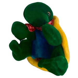 May include: A plush turtle toy with a green head, arms, and legs, a blue body, and a yellow shell. It wears a red patterned scarf around its neck. The toy is designed to look like a cartoon turtle.