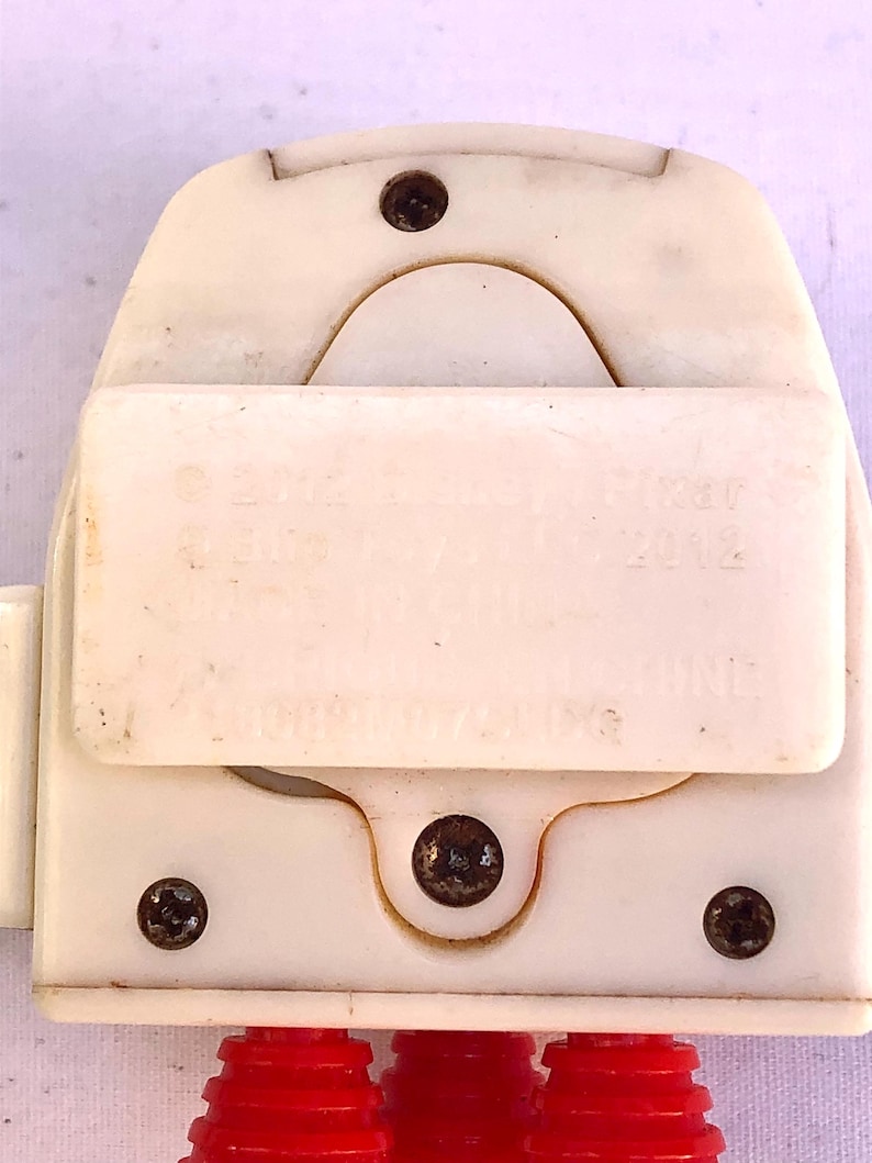 Op de afbeelding: Close-up van een wit plastic speelgoedonderdeel met drie rode, geribbelde, cilindrische verlengingen. Het onderdeel heeft een rechthoekig gedeelte met tekst en is vastgezet met vier donkere schroeven. De tekst bevat het jaar 2012.
