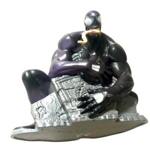 May include: A black and purple action figure of Venom, a Marvel Comics superhero, sitting on a gray, rocky base. The figure has a long, yellow tongue sticking out of its mouth.