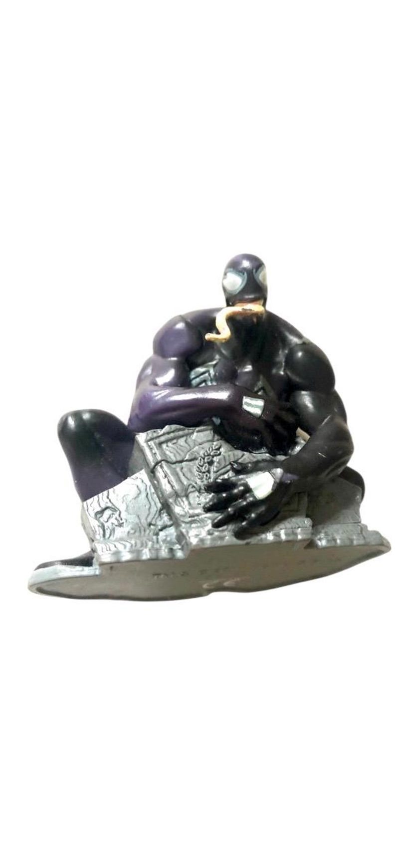 May include: A black and purple action figure of Venom, a Marvel Comics superhero, sitting on a gray, rocky base. The figure has a long, yellow tongue sticking out of its mouth.