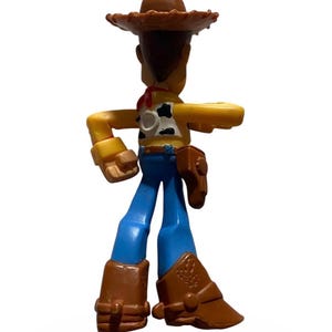 May include: A plastic toy figure of a cowboy character, wearing a brown hat, a yellow shirt with a cow pattern, blue pants, and brown boots. The figure has a red neckerchief and a brown holster. The toy is standing with one arm extended.