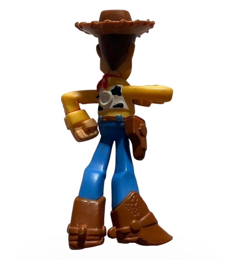 May include: A plastic toy figure of a cowboy character, wearing a brown hat, a yellow shirt with a cow pattern, blue pants, and brown boots. The figure has a red neckerchief and a brown holster. The toy is standing with one arm extended.
