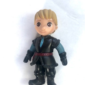 May include: A small plastic figurine of a boy with blond hair wearing a black jacket, blue sleeves, a red belt, and black boots. The figurine is from the Disney movie Frozen.