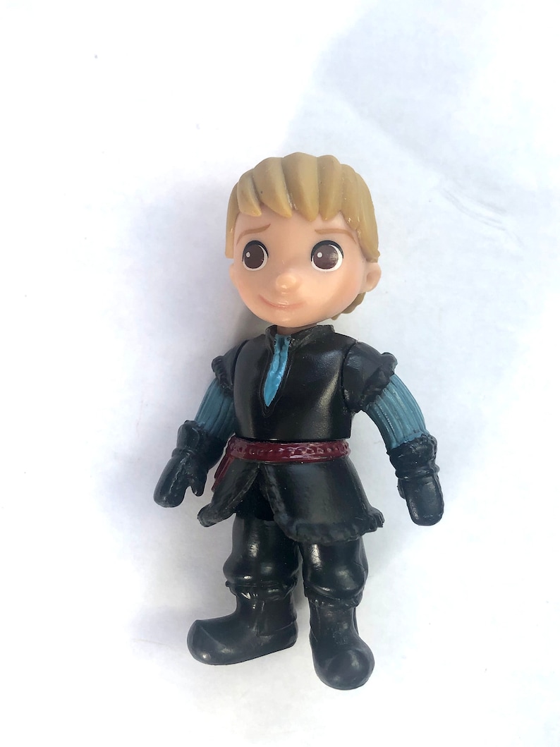 May include: A small plastic figurine of a boy with blond hair wearing a black jacket, blue sleeves, a red belt, and black boots. The figurine is from the Disney movie Frozen.