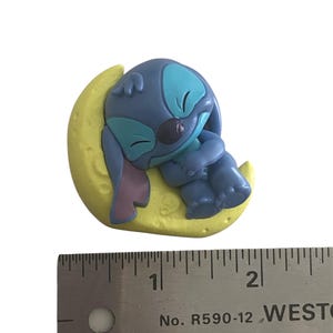 K&ouml;nnte beinhalten: Eine blaue Zeichentrickfigur, die auf einem gelben Halbmond schl&auml;ft. Die Figur hat gro&szlig;e Ohren, einen runden Kopf und umarmt ihre Knie. Der Mond ist strukturiert und hat eine kleine Herzform. Ein Lineal befindet sich neben der Figur.