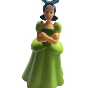 May include: A figurine of a woman in a green dress with a matching bow. The figurine has her arms crossed and a surprised expression. The dress has a full skirt and puffed sleeves. The bow is light blue.