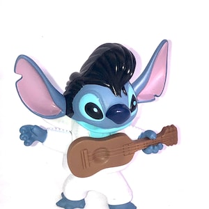 May include: Blue Stitch character with black hair, wearing a white jumpsuit and playing a brown ukulele.