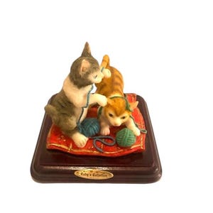 May include: A figurine of two kittens playing with balls of yarn on a red rug. The figurine is on a wooden base with the words "Ruby's Collection" inscribed on it.