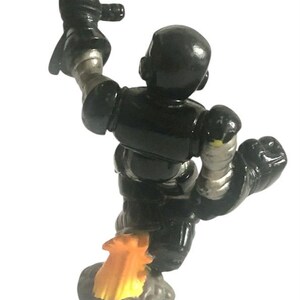 K&ouml;nnte beinhalten: Eine schwarze Spielzeugroboterfigur mit silbernen Akzenten, die mit einem Jetpack und einer Spur aus orangefarbenen Flammen fliegt. Der Roboter hat einen Arm in die Luft gestreckt.