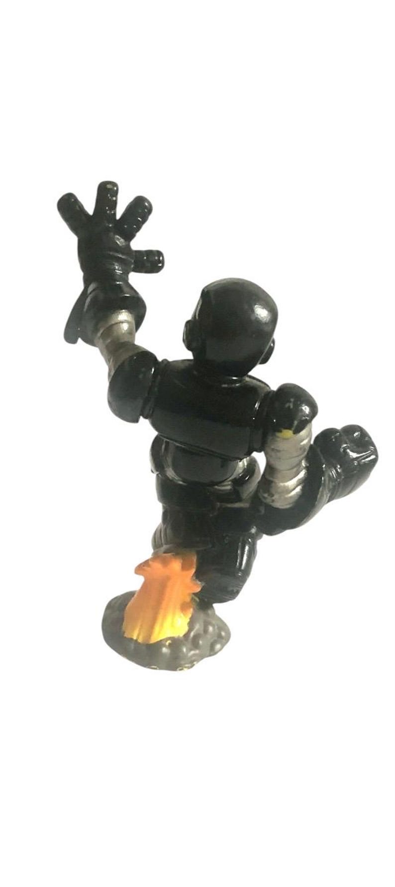 K&ouml;nnte beinhalten: Eine schwarze Spielzeugroboterfigur mit silbernen Akzenten, die mit einem Jetpack und einer Spur aus orangefarbenen Flammen fliegt. Der Roboter hat einen Arm in die Luft gestreckt.