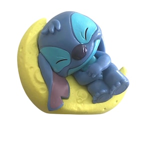 K&ouml;nnte beinhalten: Blaue Figur einer Zeichentrickfigur, die auf einem gelben Halbmond schl&auml;ft. Die Figur hat gro&szlig;e Ohren, ein hellblaues Gesicht und einen dunkelblauen K&ouml;rper. Der Mond ist strukturiert und hat ein mattes Finish.