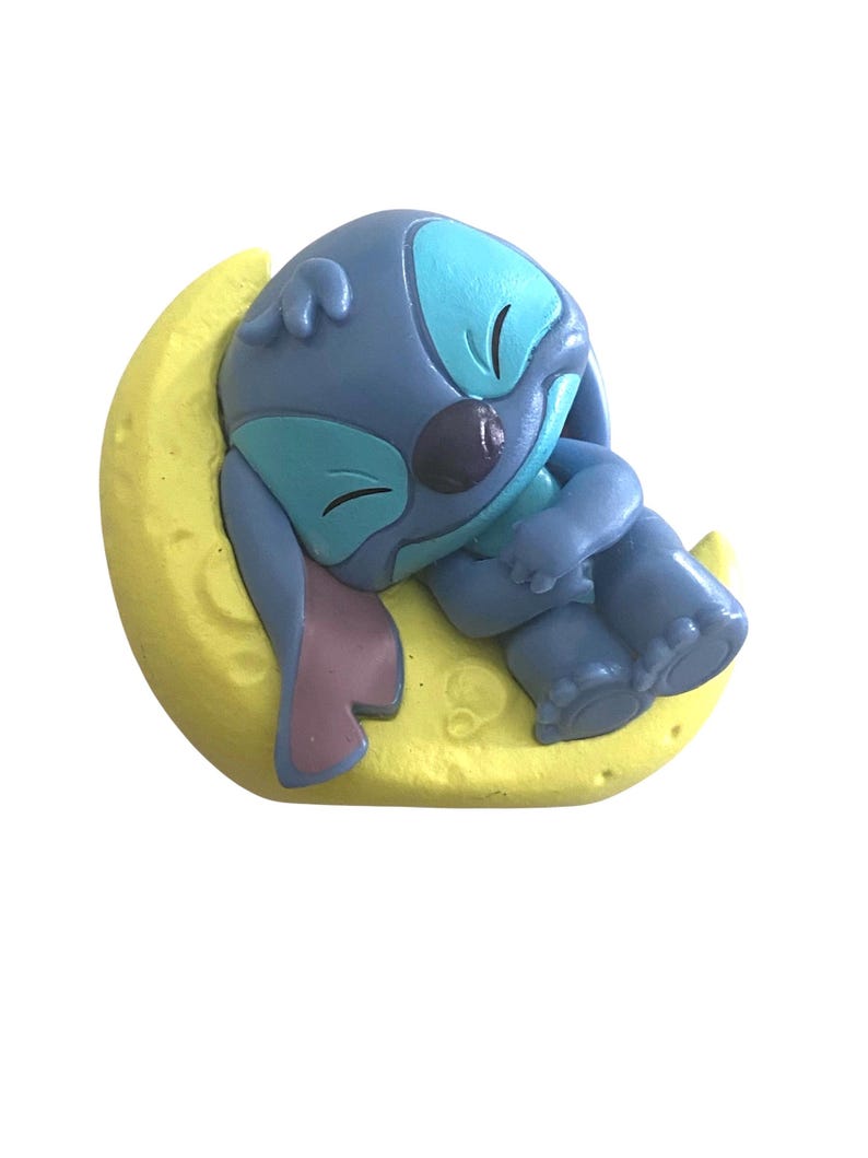 K&ouml;nnte beinhalten: Blaue Figur einer Zeichentrickfigur, die auf einem gelben Halbmond schl&auml;ft. Die Figur hat gro&szlig;e Ohren, ein hellblaues Gesicht und einen dunkelblauen K&ouml;rper. Der Mond ist strukturiert und hat ein mattes Finish.