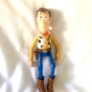 May include: A plastic toy figure of Woody from the Toy Story franchise. Woody is wearing his signature brown cowboy hat, yellow shirt with a red bandana, blue jeans, and brown boots. He has a sheriff's badge on his shirt and a holster on his hip.