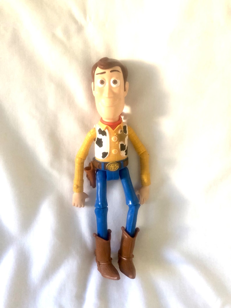 May include: A plastic toy figure of Woody from the Toy Story franchise. Woody is wearing his signature brown cowboy hat, yellow shirt with a red bandana, blue jeans, and brown boots. He has a sheriff's badge on his shirt and a holster on his hip.