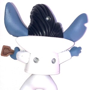 May include: Blue and white plastic figurine of a character wearing a white suit and a black hairpiece. The character is holding a brown guitar in its right hand.