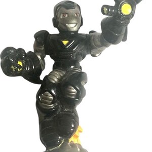 K&ouml;nnte beinhalten: Eine schwarze und silberne Actionfigur eines Superhelden, der einen schwarzen Anzug mit einem gelben Dreieck auf der Brust tr&auml;gt. Die Figur steht auf einem grauen Sockel mit einem gelben Flammenmuster. Die Figur hat ein gelbes Licht in der Hand.