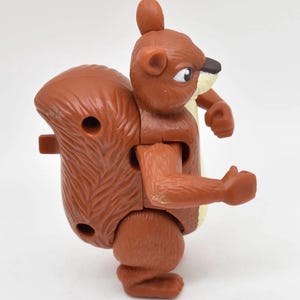 May include: A brown plastic squirrel toy with a bushy tail and articulated limbs. The toy has a detailed design, including a small black nose, white eyes, and a tuft of hair on its head. The squirrel is posed in a standing position.