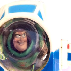 Op de afbeelding: Een close-up van een wit en blauw speelgoed met een helder, rond venster met een lachende Buzz Lightyear erin. Het speelgoed heeft een witte behuizing met blauwe accenten en een rode streep.