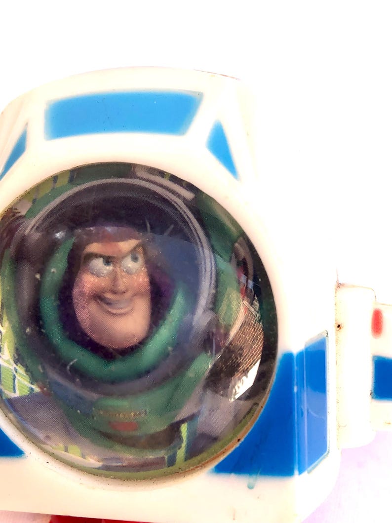 Op de afbeelding: Een close-up van een wit en blauw speelgoed met een helder, rond venster met een lachende Buzz Lightyear erin. Het speelgoed heeft een witte behuizing met blauwe accenten en een rode streep.
