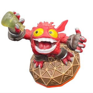 Pode incluir: Figura de ação vermelha de um monstro de desenho animado com olhos amarelos e um sorriso largo. A figura está segurando uma garrafa verde e está montada em uma base geométrica marrom e dourada. A figura tem um polegar para cima.