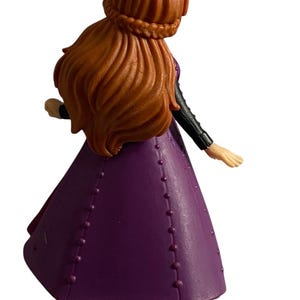 May include: A purple and black plastic doll with long brown hair wearing a purple dress with black accents. The doll is standing with one arm raised and the other arm extended to the side.