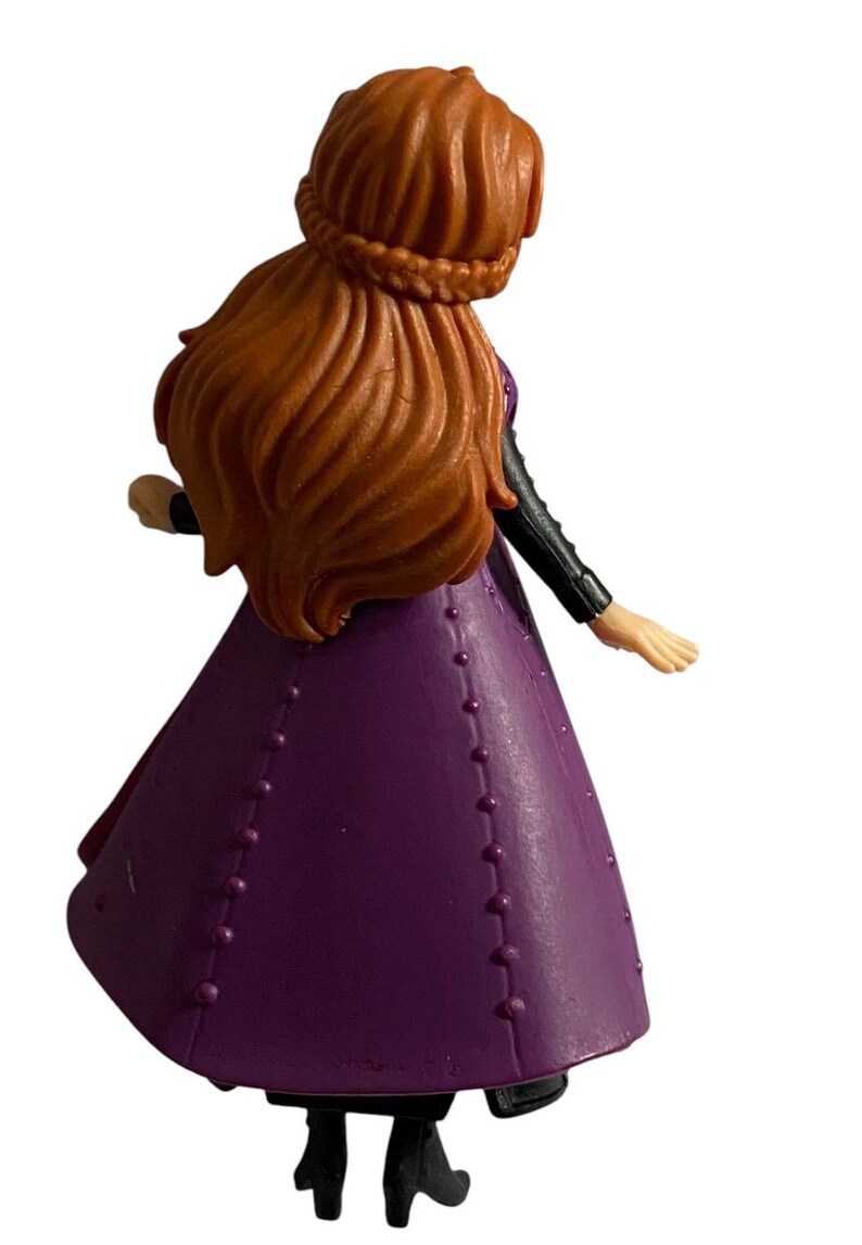 May include: A purple and black plastic doll with long brown hair wearing a purple dress with black accents. The doll is standing with one arm raised and the other arm extended to the side.