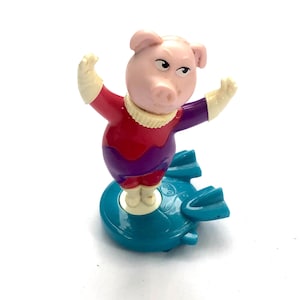 Puede incluir: Figura de plástico de un cerdo con un disfraz de superhéroe rojo y morado, con guantes blancos y un cuello crema. El cerdo está sobre una base en forma de disco de color turquesa con aletas.