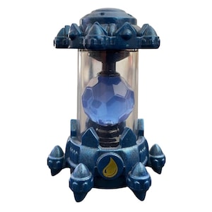 May include: A blue and silver toy with a clear cylindrical centre containing a blue, faceted sphere. The toy has a detailed, futuristic design with cone-shaped accents and a water droplet symbol.