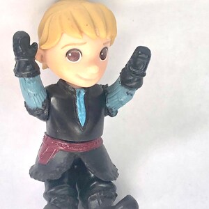 May include: A small plastic figurine of a boy with blond hair wearing a black and blue outfit with a red belt. He is holding his arms up in the air.