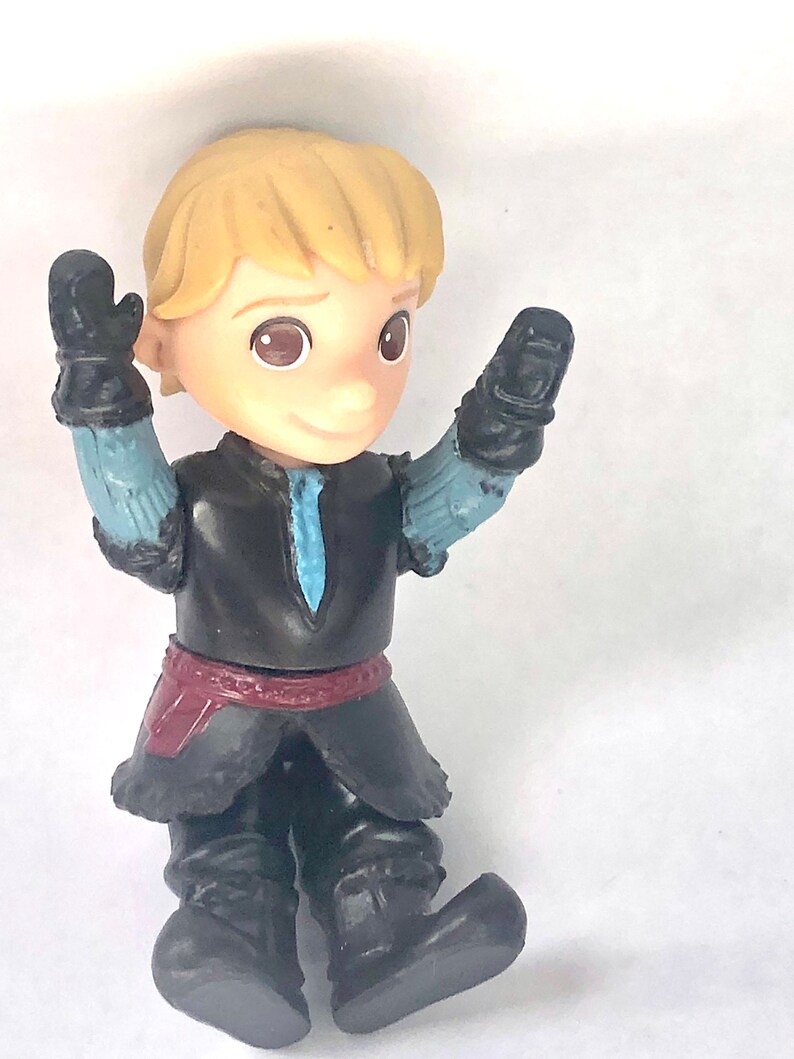 May include: A small plastic figurine of a boy with blond hair wearing a black and blue outfit with a red belt. He is holding his arms up in the air.