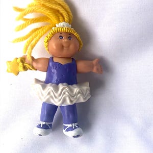May include: A small Cabbage Patch Kid doll wearing a purple and white ballerina costume with a yellow star wand. The doll has yellow yarn hair and a white crown.
